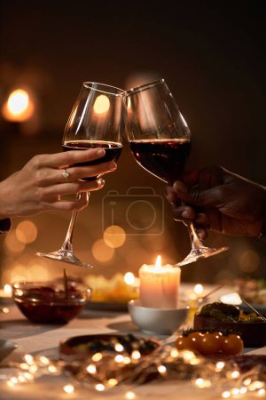 Foto de Primer plano vertical de la pareja tintineando copas de vino en la mesa de la cena con luces de Navidad en el fondo - Imagen libre de derechos