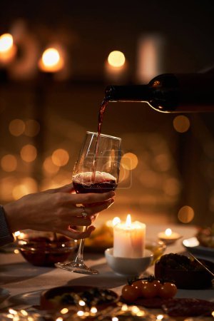 Foto de Primer plano de vino tinto vertiendo en vidrio en la mesa de la cena con velas y luces de Navidad en el fondo - Imagen libre de derechos