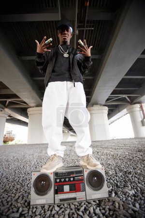 Photo for Long shot of cool rapper in trendy attire standing on top of old tape recorder in urban environment under bridge during performance - Royalty Free Image