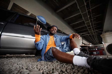 Happy young African American male zoomer throwing rugby ball while sitting on ground against silver car and listening to old tape recorder