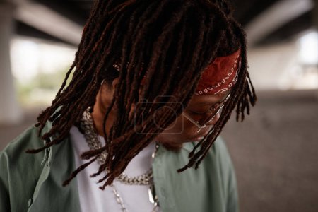 Téléchargez les photos : Serrures de lecture de jeune artiste afro-américain hip hop élégant avec bandeau sur la tête et des lunettes de soleil debout devant la caméra - en image libre de droit