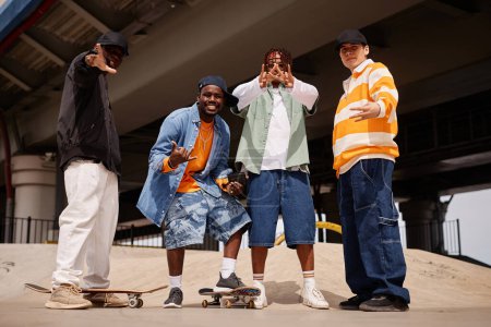 Foto de Cuatro jóvenes zoómeros masculinos interculturales felices en ropa casual con estilo mostrando varios gestos y mirando a la cámara al aire libre - Imagen libre de derechos