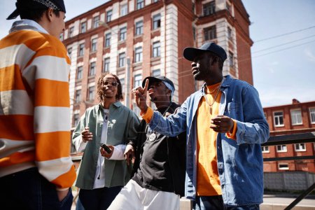 Gruppe junger interkultureller Männer in stylischer Kleidung, die im Freien Hip-Hop spielen, während sie vor der Kamera vor modernen Gebäuden stehen