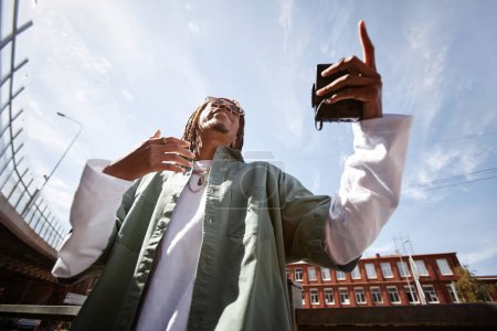 Téléchargez les photos : Vue d'en bas de jeune homme afro-américain excité avec walkman à la main exécutant hip hop contre ciel bleu en milieu urbain - en image libre de droit