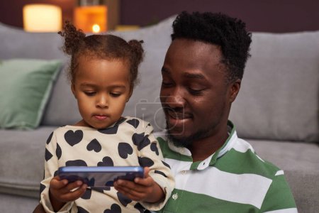 Foto de Retrato del joven padre negro y su hija usando un teléfono inteligente juntos para el tiempo de pantalla supervisado - Imagen libre de derechos