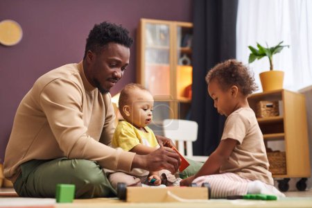 Photo for Side view portrait of caring Black father playing with two children sitting on floor at home and enjoying fatherhood - Royalty Free Image