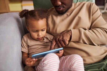 Gros plan du père enseignant la petite fille noire à l'aide d'un smartphone tout en étant assis ensemble sur le canapé à la maison, copiez l'espace