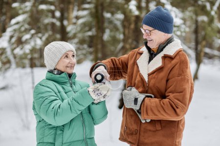 Smiling senior man pouring hot tea for wife when they are stand in winter forest