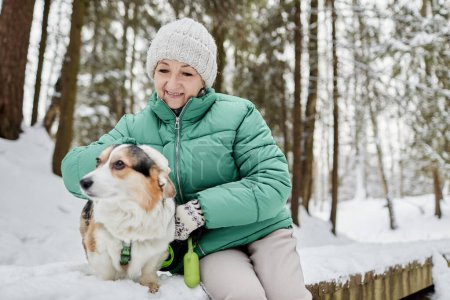 Smiling senior woman patting her corgi dog when walk in winter park