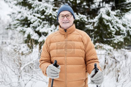 Portrait of joyful senior man in warm clothes skiing in park on sunny winter