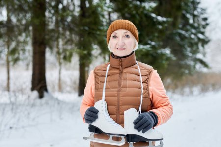 Portrait of positive smile senior woman in warm clothes stand in forest with ice skates