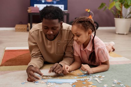 Photo for Portrait of Black little girl solving jigsaw puzzle while laying on floor with father and playing together at home - Royalty Free Image