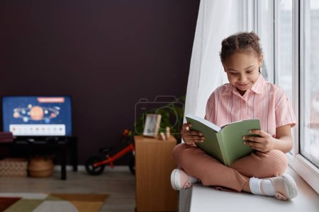 Foto de Retrato de cuerpo entero de una linda niña negra sentada en la ventana y leyendo el libro en el interior de la habitación rosa, espacio para copiar - Imagen libre de derechos