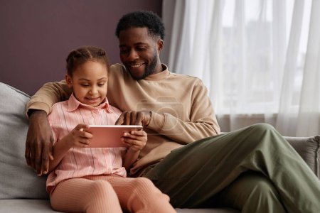 Foto de Retrato de padre e hija negros usando tableta digital juntos sentados en el sofá en casa y disfrutando del tiempo en familia, espacio para copiar - Imagen libre de derechos