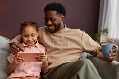 Portrait de heureux père noir et petite fille en utilisant la tablette numérique ensemble assis sur le canapé à la maison et profiter du temps de la famille, copier l'espace