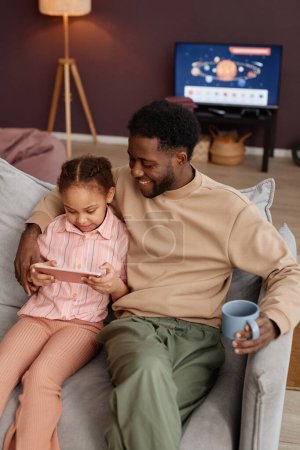 Foto de Retrato vertical de un hombre negro sonriente con una hija pequeña usando la tableta juntos sentados en el sofá en casa - Imagen libre de derechos