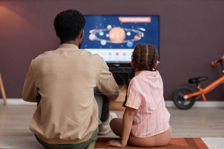 Téléchargez les photos : Vue arrière du père et de la fille regardant des vidéos éducatives sur l'espace ensemble assis sur le sol devant la télévision - en image libre de droit