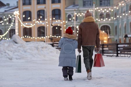 Téléchargez les photos : Pleine longueur vue arrière du père et de l'enfant marchant à l'extérieur en hiver tenant des sacs à provisions colorés, espace de copie - en image libre de droit