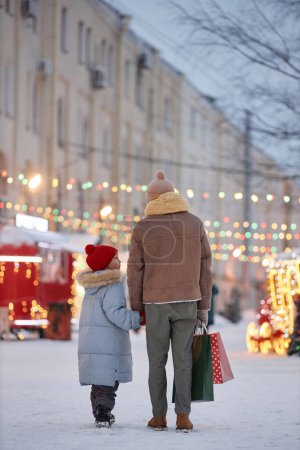 Foto de Vista posterior vertical del padre y el niño tomados de la mano caminando en el parque de invierno decorado para Navidad en la ciudad - Imagen libre de derechos