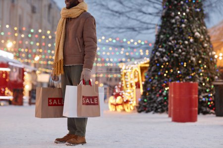 Foto de Recortado tiro de sonriente hombre negro sosteniendo bolsas de compras al aire libre en invierno con decoraciones de Navidad, espacio de copia - Imagen libre de derechos