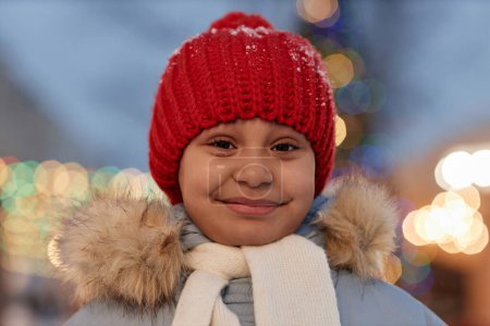 Foto de Primer plano retrato de linda chica negra sonriendo a la cámara con chaqueta de invierno y sombrero de punto rojo al aire libre con luces en el fondo - Imagen libre de derechos