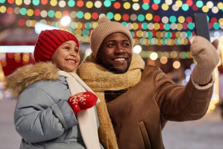 Téléchargez les photos : Homme et fille noirs prenant selfie photo en plein air ensemble dans le marché de Noël et souriant, espace de copie - en image libre de droit