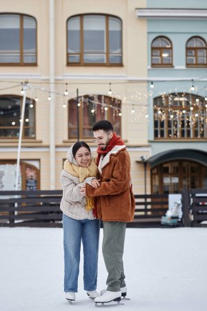 Foto de Retrato vertical de larga duración de una pareja joven patinando juntos en una pista de patinaje al aire libre y divirtiéndose en invierno - Imagen libre de derechos