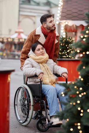 Photo for Vertical full length portrait of young man assisting girlfriend with disability outdoors at Christmas market - Royalty Free Image