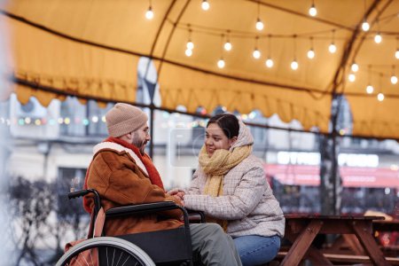Foto de Retrato de vista lateral de una joven sonriente cogida de la mano con un hombre en silla de ruedas disfrutando de una cita al aire libre en invierno - Imagen libre de derechos