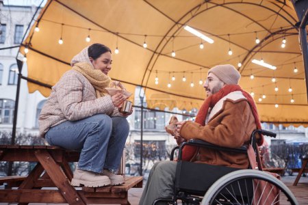 Foto de Retrato de hombre en silla de ruedas compartiendo bebidas calientes con una mujer joven disfrutando de una cita en la ciudad de invierno - Imagen libre de derechos