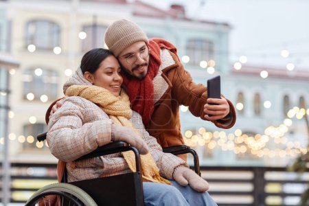 Foto de Retrato de pareja joven con una mujer en silla de ruedas tomando fotos de selfies al aire libre en invierno y usando un teléfono inteligente - Imagen libre de derechos