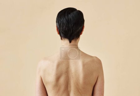 Foto de Vista trasera de una mujer adulta irreconocible con la espalda desnuda de pie sobre un fondo beige mínimo, espacio para copiar - Imagen libre de derechos