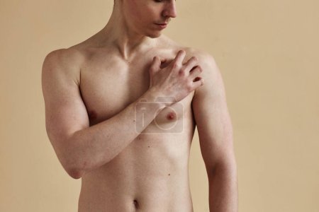 Téléchargez les photos : Gros plan de jeune homme torse nu qui gratte la peau debout sur un fond pâle, espace de copie - en image libre de droit