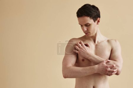 Téléchargez les photos : Portrait taille haute minime de jeune homme torse nu qui gratte la peau sur fond beige, espace de copie - en image libre de droit
