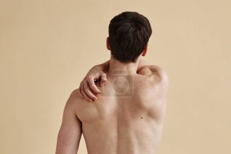 Foto de Vista trasera mínima del hombre rascarse la espalda desnuda de pie contra el fondo beige, espacio de copia - Imagen libre de derechos