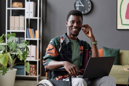 Foto de Retrato de un joven negro usando una silla de ruedas sonriendo a la cámara y sosteniendo el portátil en la oficina, espacio para copiar - Imagen libre de derechos