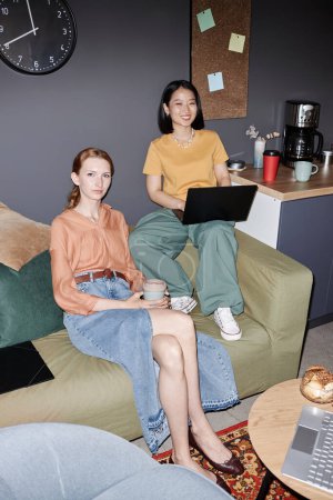Foto de Retrato vertical de longitud completa de dos mujeres jóvenes modernas sonriendo a la cámara disfrutando del trabajo en el salón de oficina con flash - Imagen libre de derechos
