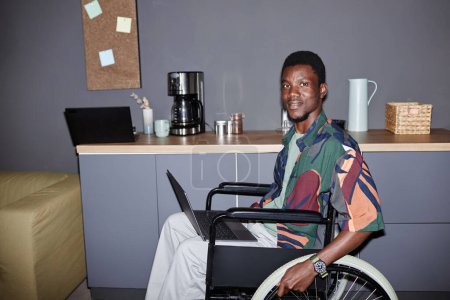 Portrait of one young Black man using wheelchair and smiling at camera in modern office lounge, shot with flash, copy space