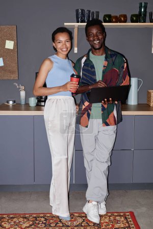 Vertikales Ganzkörperporträt zweier schwarzer junger Menschen, die an einer Kaffeestation in der Bürolounge stehen und mit Blitz lächeln