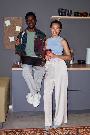 Vertikales Ganzkörperporträt zweier schwarzes junges Paar, das an einer Kaffeestation in der Bürolounge steht und in die Kamera lächelt