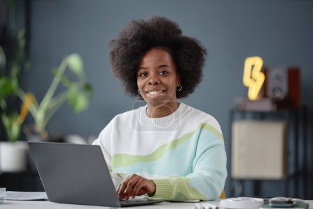 Foto de Retrato mínimo de una mujer afroamericana sonriente con cabello rizado natural usando una computadora portátil en la oficina y mirando el espacio de copia de la cámara - Imagen libre de derechos