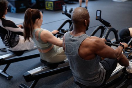 Photo for Back view of people using rowing machine during strength training in gym with muscular man in foreground - Royalty Free Image