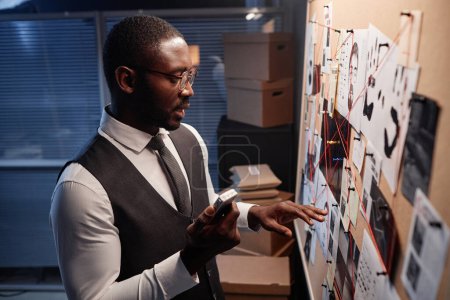 Side view portrait of Black male detective holding voice recorder standing by evidence board, copy space