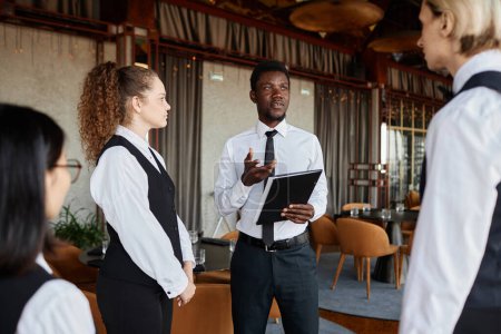 Téléchargez les photos : Portrait de jeune homme noir parlant à des serveurs portant des uniformes classiques lors d'une réunion du personnel dans un restaurant - en image libre de droit