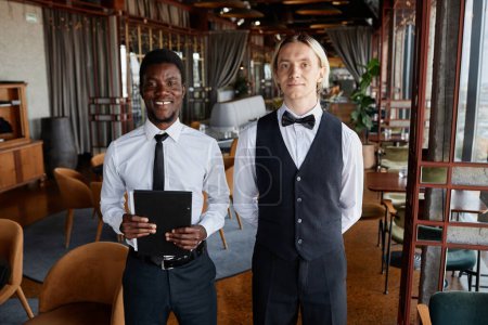 Téléchargez les photos : Portrait taille haute de deux jeunes hommes comme personnel de restaurant de luxe portant des uniformes classiques en noir et blanc et regardant la caméra, l'espace de copie - en image libre de droit