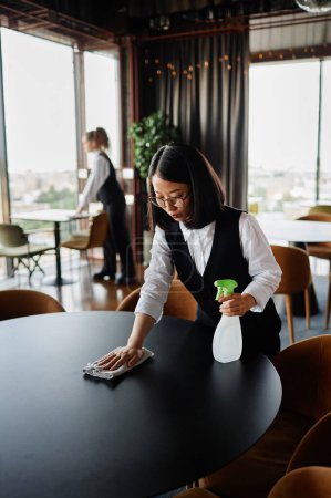 Téléchargez les photos : Portrait vertical de la jeune femme asiatique comme serveur nettoyage table à manger dans le restaurant se préparant pour l'ouverture - en image libre de droit