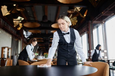 Retrato de hombre joven como camarero vestido con uniforme clásico blanco y negro limpiando mesas en un restaurante de lujo, espacio para copiar