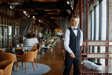 Téléchargez les photos : Portrait de jeune homme élégant portant smoking nettoyage uniforme avec plumeau dans l'intérieur du restaurant de luxe, espace de copie - en image libre de droit