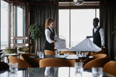 Photo for Side view portrait of two elegant waiters setting tables in luxury restaurant and holding white tablecloth, copy space - Royalty Free Image