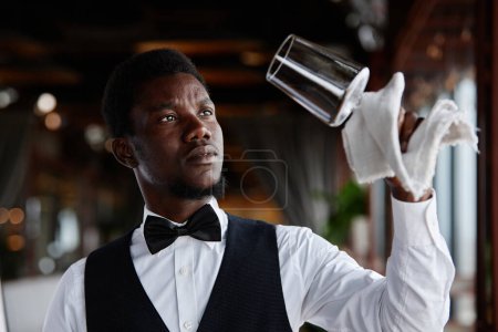 Téléchargez les photos : Portrait de jeune homme noir comme élégant serveur verres à polir dans un restaurant de luxe, espace de copie - en image libre de droit